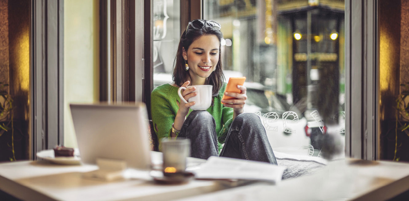 Young woman sitting in a coffee shop while using her phone and laptop.